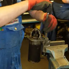 Mechanik w niebieskim kombinezonie i czerwonych rękawicach dokonuje wymiany szczotek w rozruszniku, który jest zamocowany w imadle.