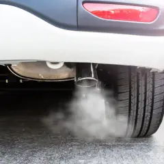 Białe auto pluje wodą z wydechu, tworząc mgiełkę na asfalcie. Widać fragment opony i tylną lampę.