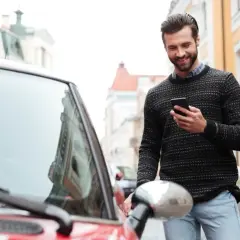 Uśmiechnięty mężczyzna z telefonem w ręku stoi obok czerwonego samochodu na ulicy.
