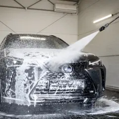 Czarny Lexus jest obficie spieniony podczas mycia na myjni bezdotykowej. Widać strumień wody z pianą, który opływa maskę i grill.