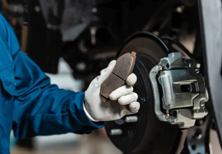 Mechanik w niebieskim kombinezonie trzyma zużyty klocek hamulcowy, zastanawiając się, ile kosztuje wymiana zacisku hamulcowego.