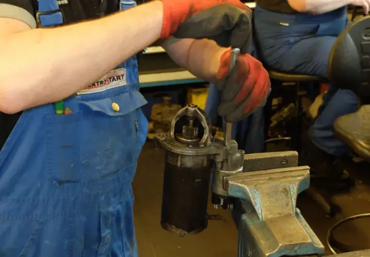 Mechanik w niebieskim kombinezonie i czerwonych rękawicach dokonuje wymiany szczotek w rozruszniku, który jest zamocowany w imadle.