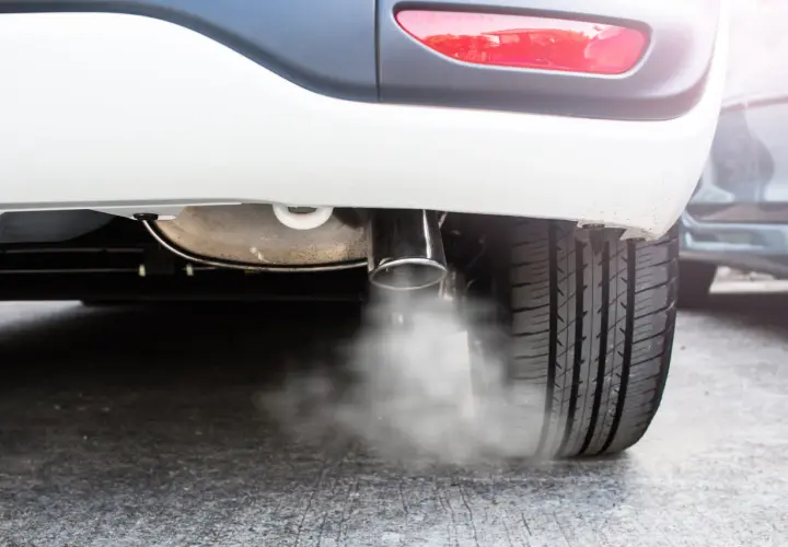 Białe auto pluje wodą z wydechu, tworząc mgiełkę na asfalcie. Widać fragment opony i tylną lampę.