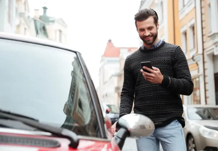 Uśmiechnięty mężczyzna z telefonem w ręku stoi obok czerwonego samochodu na ulicy.