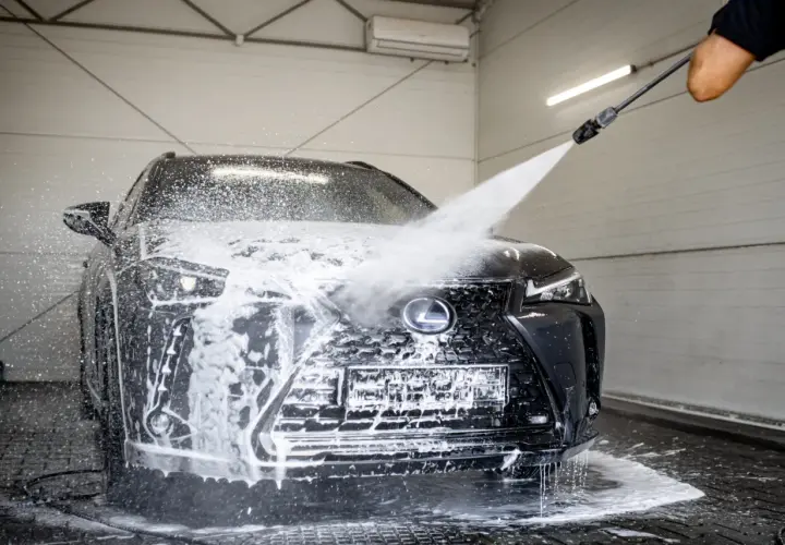 Czarny Lexus jest obficie spieniony podczas mycia na myjni bezdotykowej. Widać strumień wody z pianą, który opływa maskę i grill.