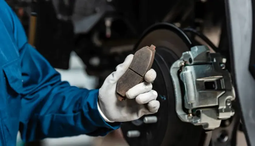 Mechanik w niebieskim kombinezonie trzyma zużyty klocek hamulcowy, zastanawiając się, ile kosztuje wymiana zacisku hamulcowego.