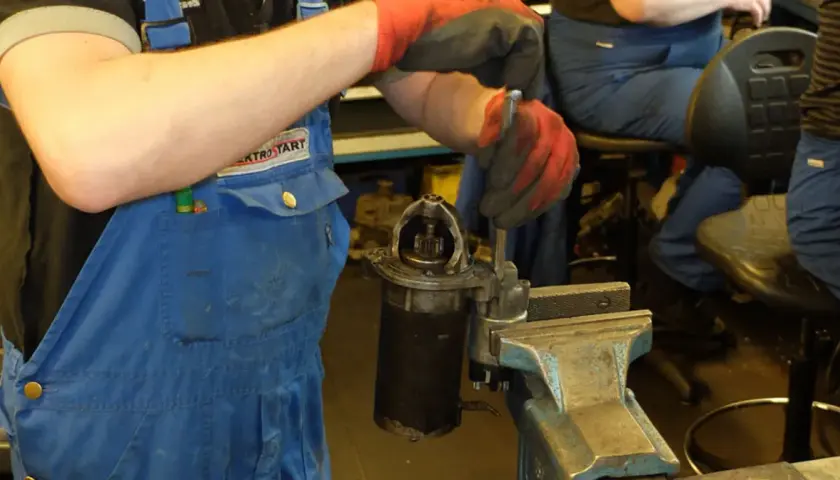 Mechanik w niebieskim kombinezonie i czerwonych rękawicach dokonuje wymiany szczotek w rozruszniku, który jest zamocowany w imadle.