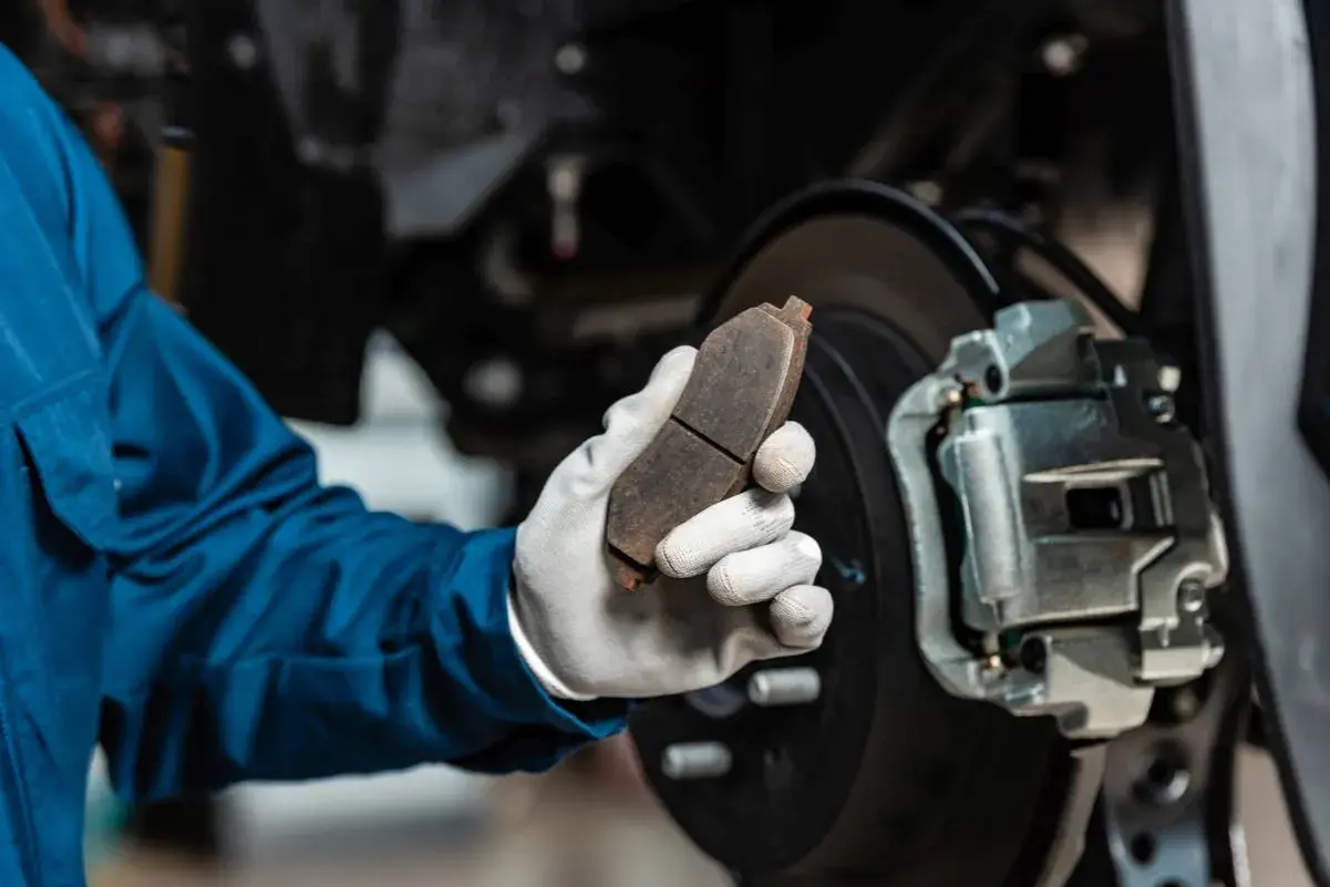 Mechanik w niebieskim kombinezonie trzyma zużyty klocek hamulcowy, zastanawiając się, ile kosztuje wymiana zacisku hamulcowego.