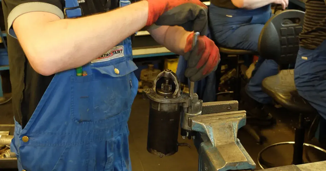 Mechanik w niebieskim kombinezonie i czerwonych rękawicach dokonuje wymiany szczotek w rozruszniku, który jest zamocowany w imadle.