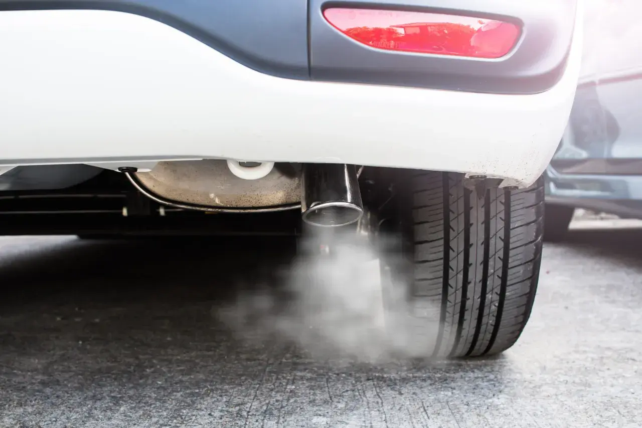 Białe auto pluje wodą z wydechu, tworząc mgiełkę na asfalcie. Widać fragment opony i tylną lampę.