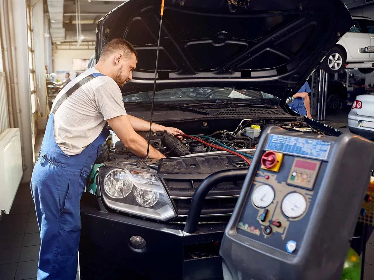 Mechanik naprawia klimatyzację w samochodzie, słychać syczenie z klimatyzacji. Urządzenie diagnostyczne stoi obok.