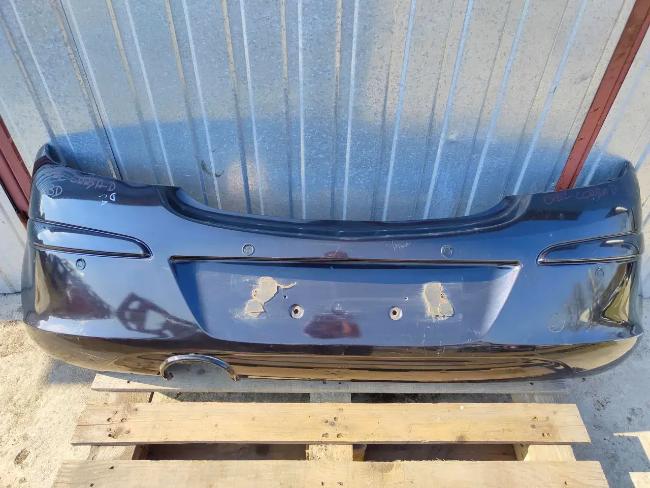 Ciemny zderzak Opel Corsa D z widocznymi śladami użytkowania, leżący na drewnianej palecie.