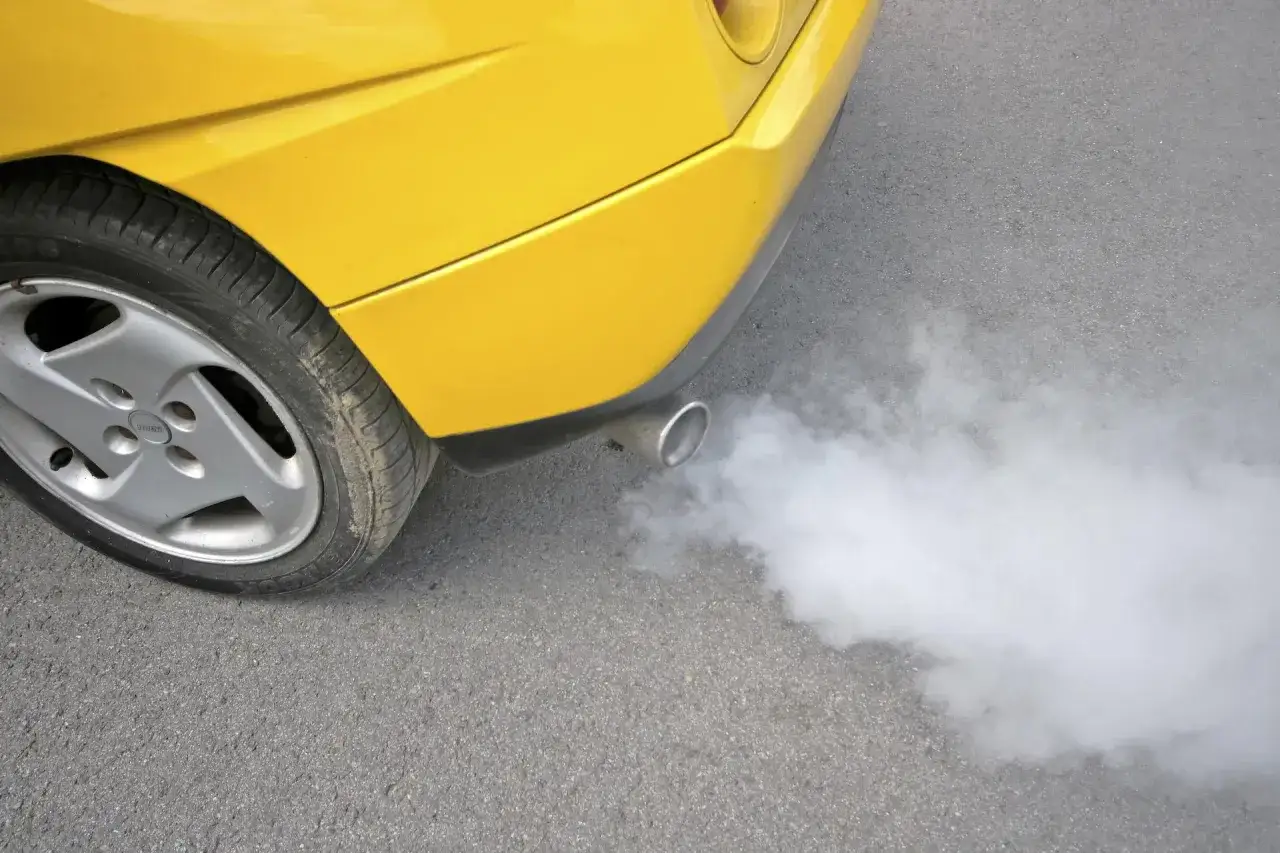 Żółte auto pluje wodą z wydechu, tworząc białą chmurę pary na szarym asfalcie.