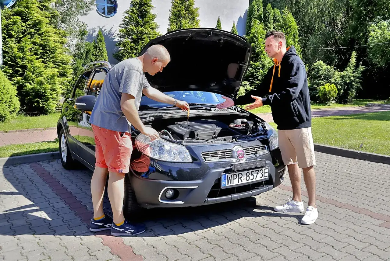 Dwóch mężczyzn ogląda silnik czarnego Fiata. Jeden sprawdza poziom oleju, drugi gestykuluje. Wskazówka, jak negocjować cenę samochodu używanego.
