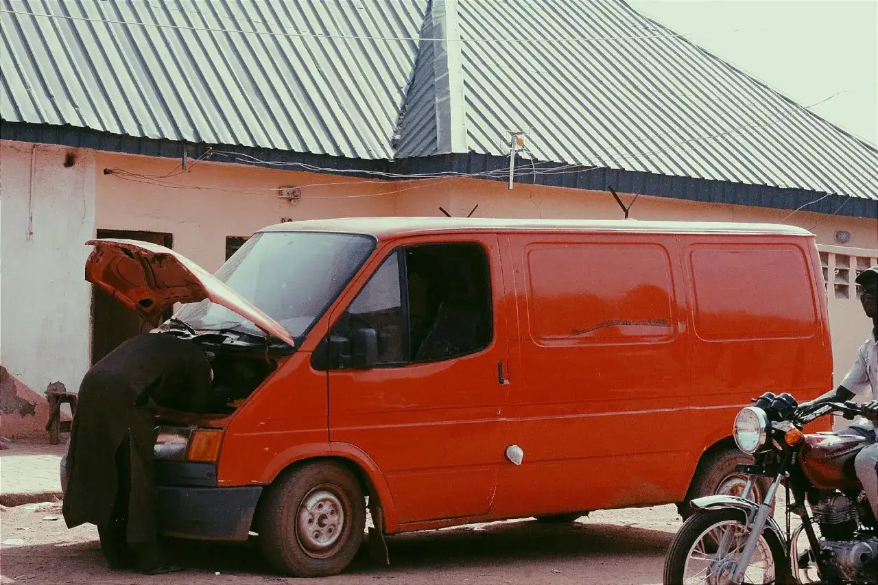 Mechanik pochyla się nad silnikiem pomarańczowego vana, by sprawdzić czujnik ciśnienia w kolektorze ssącym. Obok stoi motocykl.