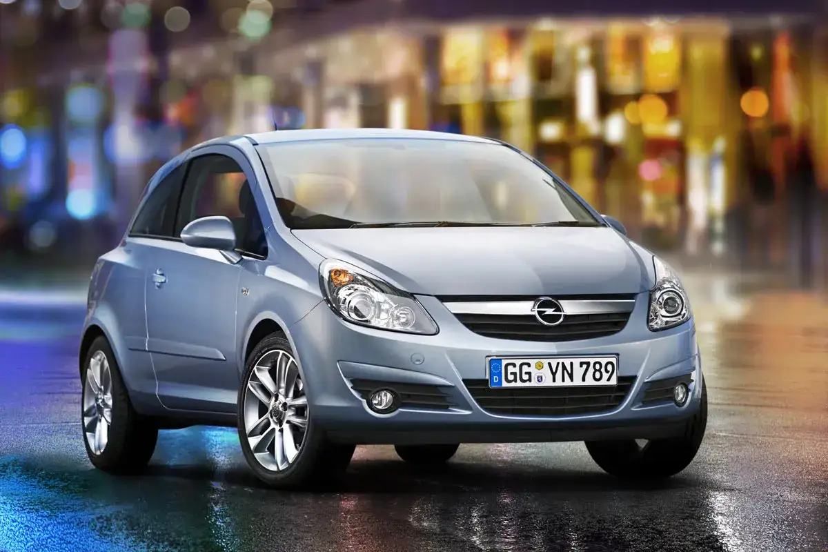 Błękitny Opel Corsa D z błyszczącym błotnikiem, zaparkowany na mokrej ulicy nocą, z rozmytymi światłami miasta w tle.